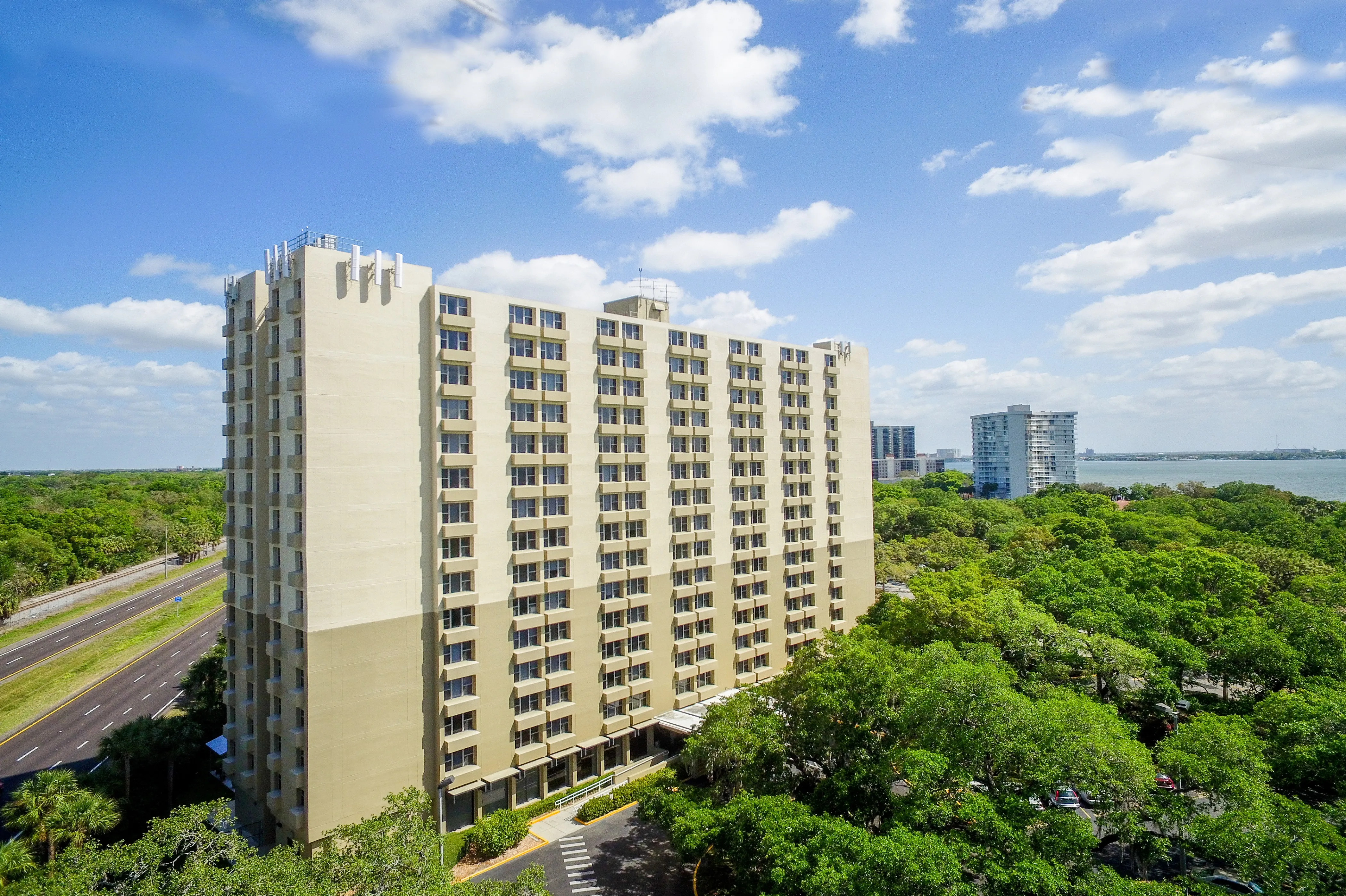 A beige high-rise apartment building surrounded by lush trees with Hillsborough Bay in the background.